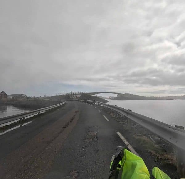 Straße am Wasser bei bewölktem Himmel, mit Brücke in der Ferne und Motorradhelm im Vordergrund.