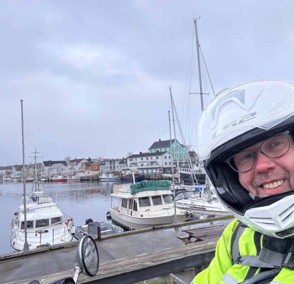 Person mit Helm lächelt am Hafen; im Hintergrund Boote und Gebäude bei bewölktem Himmel.