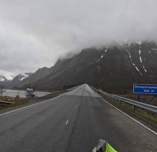 Leere Landstraße führt auf bewölkte Berge zu, mit einem See links und einem blauen Straßenschild rechts.