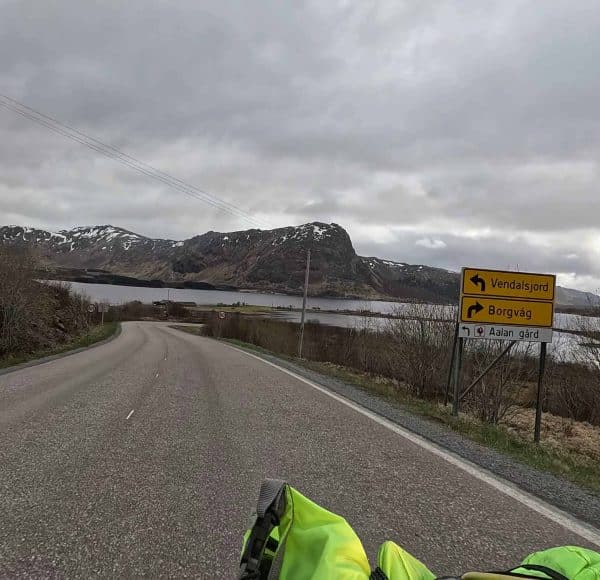 Landstraße mit Kurve, Straßenschild rechts, Berge und See im Hintergrund, bewölkter Himmel, Motorradfahrer unten im Bild.