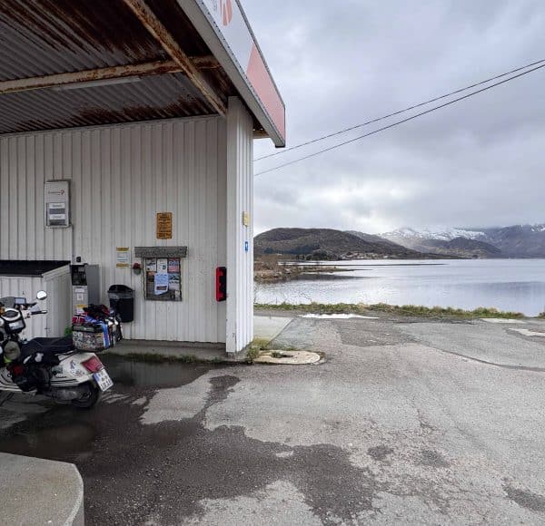 Tankstelle am Seeufer mit einem Motorrad vorne, bewölkter Himmel und Berge im Hintergrund.