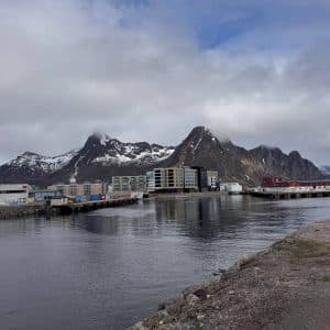 Küstenstadt mit modernen Gebäuden am Wasser, im Hintergrund schneebedeckte Berge und bewölkter Himmel.