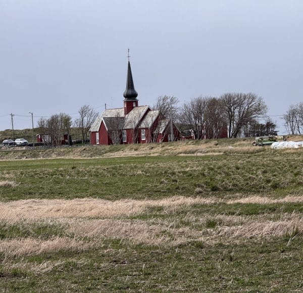 Kirche mit hohem Turm steht auf einem weiten, grasigen Feld unter grauem Himmel, umgeben von Bäumen und wenigen Gebäuden.
