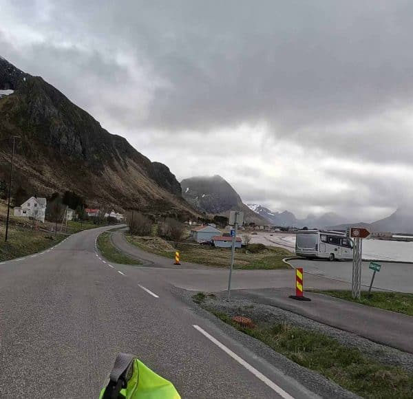 Straße führt durch ein kleines Dorf, umgeben von Bergen und bewölktem Himmel.