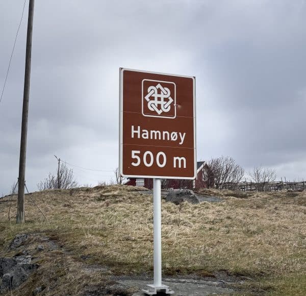 Braunes Straßenschild mit der Aufschrift „Hamnøy 500 m“, aufgestellt auf einer Wiese unter bewölktem Himmel.