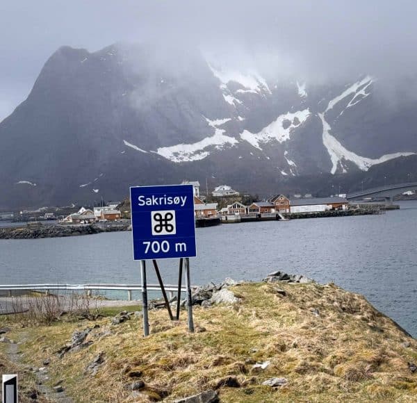 Blaues Straßenschild mit „Sakrisøy 700 m“ vor Wasser, Dorf und schneebedecktem Berg im Hintergrund.