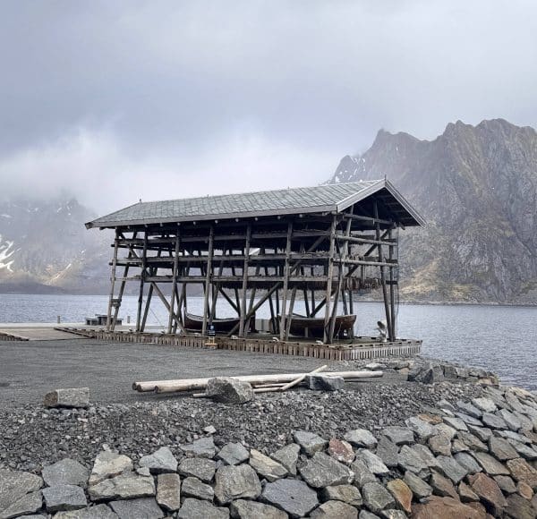 Holzgerüst mit Stockfisch am Reine Fjord, umgeben von Bergen und bewölktem Himmel, mit Steinen im Vordergrund.