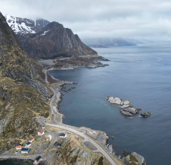 Drohnenaufnahme der Küstenlandschaft östlichen Teils des Reine Fjord mit kurvenreicher Straße entlang felsiger Berge, das Meer mit kleinen Felsen, bewölkter Himmel und vereinzelt Schnee auf den Gipfeln.