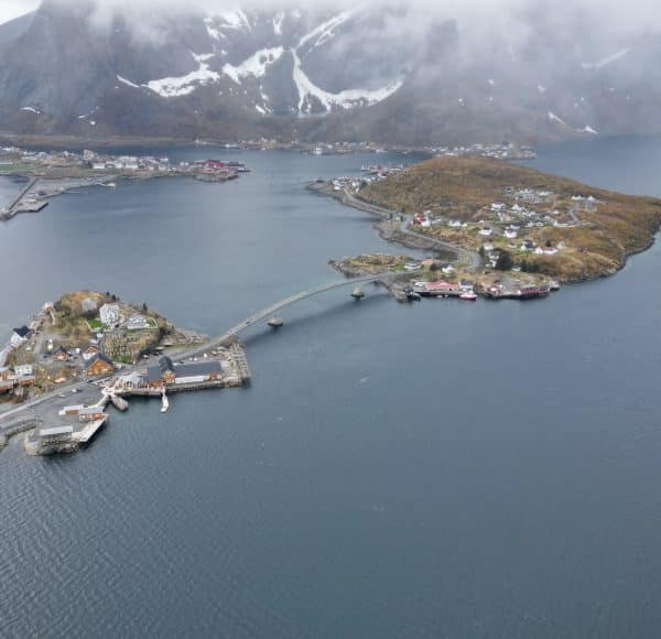 Luftaufnahme einer kleinen Inselstadt mit Brücke, umgeben von Wasser und Bergen im Hintergrund, teils schneebedeckt.