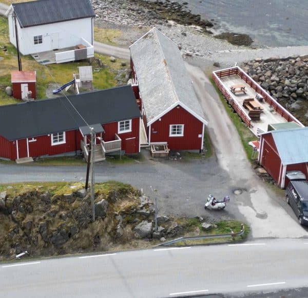 Mehrere rote Holzhäuser stehen nahe einer Küste, mit Straßen, felsigem Gelände und dem Meer im Hintergrund.