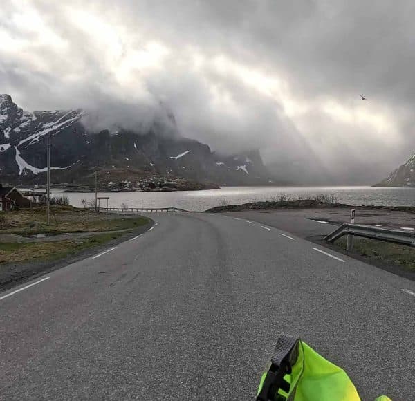 Leere Straße am See, Berge im Hintergrund, bewölkter Himmel, etwas Schnee, neongrüner Motorradhelm vorne im Bild.