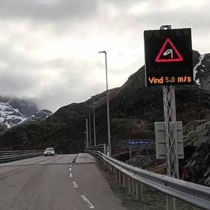 Straße mit elektronischem Warnschild, das vor Steinschlag und Wind bis 18 m/s warnt, bergige Landschaft im Hintergrund.