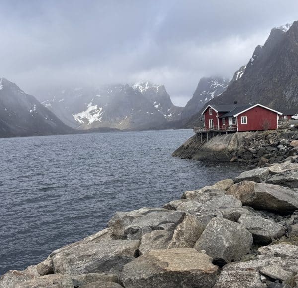 Rotes Haus an felsiger Küste mit Blick auf einen Fjord, umgeben von schneebedeckten Bergen unter bewölktem Himmel.