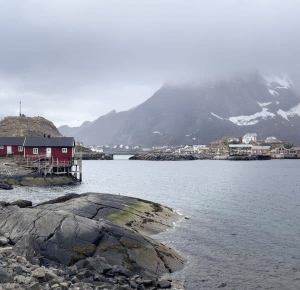 Rotes Haus am Wasser, felsige Küste, Berge im Hintergrund und bewölkter Himmel; ruhige nordische Landschaft im Nebel.