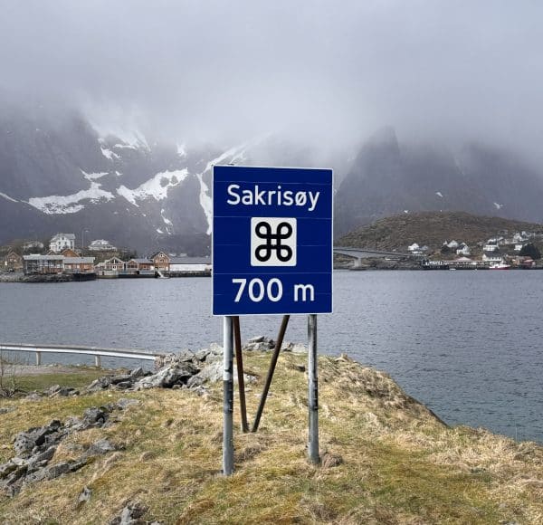 Blaues Verkehrsschild mit „Sehenswürdigkeit 700 m Sakrisoy“, Symbol, im Vordergrund, im Hintergrund Berge, Reine-Fjord und einige Gebäude, bewölktes Wetter.