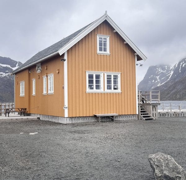 Gelbes Holzhaus steht vor Bergen und See an einem bewölkten Tag.