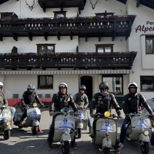 Sechs Erwachsene mit Helmen sitzen auf klassischen Rollern vor einer Pension mit Holzbalkonen und Blumen, daneben Autos und Wandmalerei.