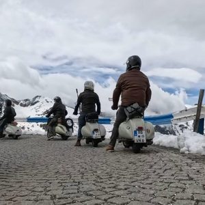 Vier Personen fahren mit der Vespa auf einer gepflasterten Bergstraße von der Edelweißspitze, umgeben von Schnee und Bergen, unter wolkigem Himmel.
