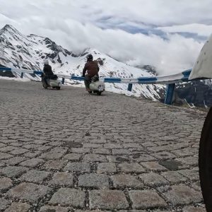 Zwei Menschen fahren auf Motorrollern eine gepflasterte Bergstraße entlang, umgeben von schneebedeckten Gipfeln und bewölktem Himmel.