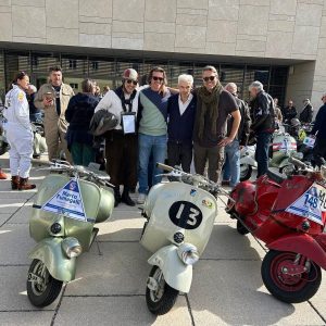 Eine Gruppe von Menschen posiert vor klassischen Vespa-Rollern bei einer Veranstaltung im Freien, weitere Teilnehmende und Roller im Hintergrund.