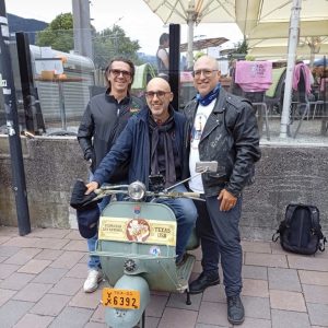Drei Männer posieren lächelnd mit einer Vintage-Vespa auf einem Bürgersteig vor einem Straßencafé mit Sonnenschirmen und bunten Decken.
