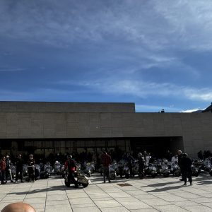 Viele Menschen und zahlreiche Motorroller versammeln sich bei sonnigem Wetter auf einem großen, gepflasterten Platz vor einem modernen Gebäude.
