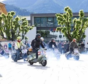 Mehrere Menschen fahren Motorroller auf einem hellen Platz, Zuschauer stehen im Hintergrund, Bäume und Berge im Hintergrund, sonniges Wetter.