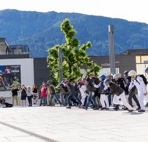 Menschen in Raumanzügen rennen auf einem Platz, im Hintergrund Berge, Bäume und staunende Zuschauer.