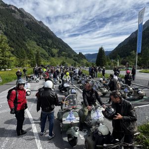 Viele Menschen mit Motorrollern versammeln sich auf einem Parkplatz in einem grünen Tal, umgeben von Bergen und bewölktem Himmel.