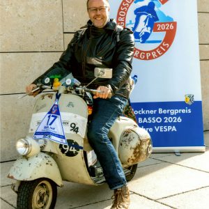 Mann mit Lederjacke sitzt auf alter Vespa vor einem Poster für den Grossglockner Bergpreis 2026.