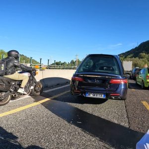 Motorradfahrer und mehrere Autos stehen im Stau auf einer Autobahn bei sonnigem Wetter, im Hintergrund Bäume und blauer Himmel.