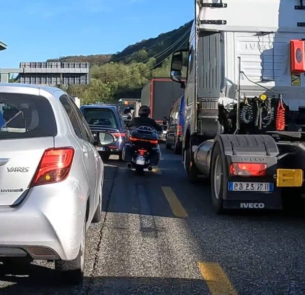 Autos, LKW stehen auf der italienischen Autobahn im Stau, ein Motorrad-Fahrer schlängelt sich zwischen den Autos in der Mitte beider Fahrspuren durch.