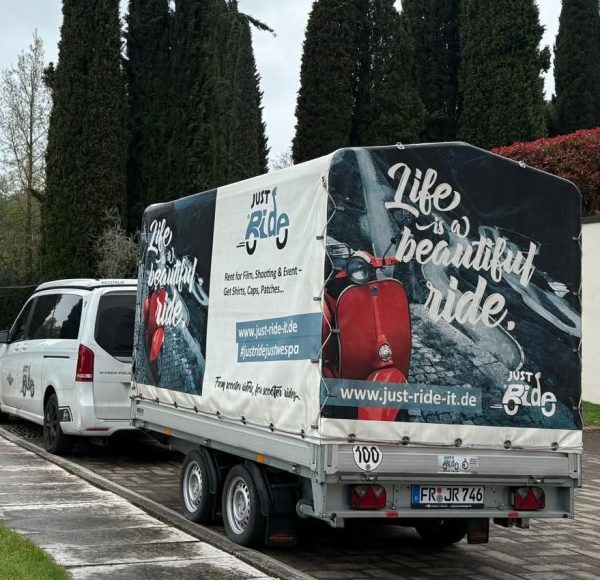 Weißer Lieferwagen mit Anhänger, darauf Werbung mit dem Spruch „Life is a beautiful ride“, neben einer Straße und hohen Bäumen.