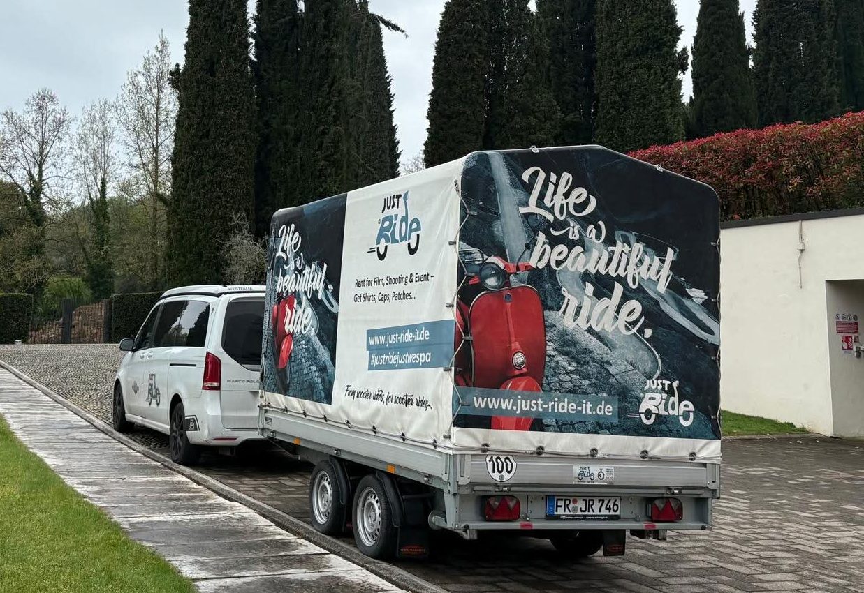 Weißer Lieferwagen mit Anhänger, darauf Werbung mit dem Spruch „Life is a beautiful ride“, neben einer Straße und hohen Bäumen.