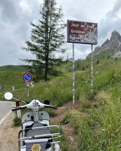 Ein Motorroller steht vor einem mit Aufklebern bedeckten Passschild am Straßenrand in den Bergen, umgeben von grünem Gras und felsigen Gipfeln.