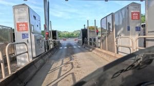 Mautstation mit Schranke und roten Ampeln, Automaten auf beiden Seiten, Landschaft im Hintergrund, klare Himmel.