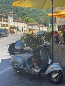 Blaue Vintage-Vespa mit Helm, Croissant und Kaffee auf Sitz, unter gelbem Sonnenschirm, im Straßencafé eines malerischen italienischen Dorfes mit grünen Hügeln im Hintergrund.