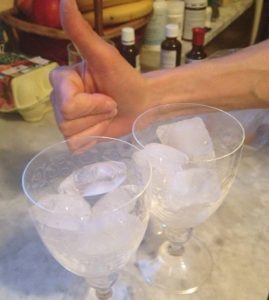 Zwei Weingläser mit Eiswürfeln auf einem Tisch, im Hintergrund zeigt eine Hand den Daumen nach oben.