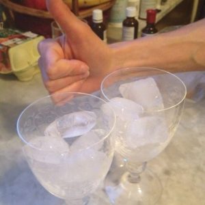 Zwei Weingläser mit Eiswürfeln auf einem Tisch, im Hintergrund zeigt eine Hand den Daumen nach oben.