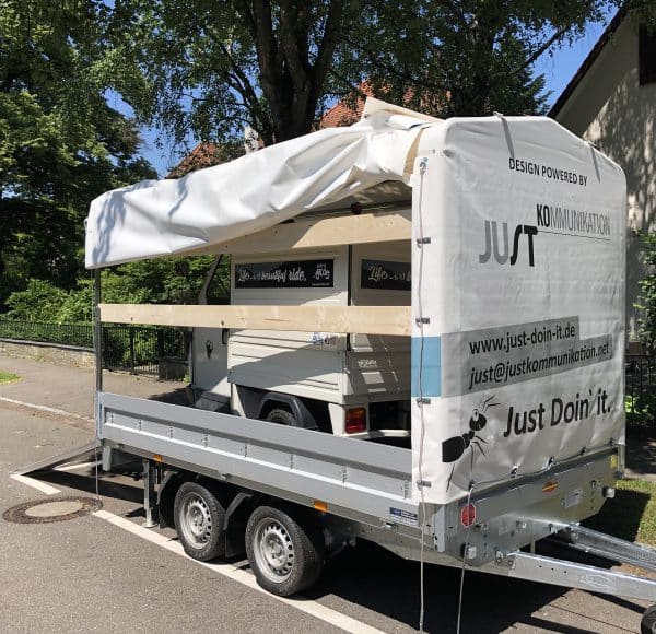 Ein Anhänger mit Plane und Werbeaufschrift „Just Do it“, darauf ein kleinerer Transporter, im Hintergrund Bäume und Haus, sonniges Wetter.