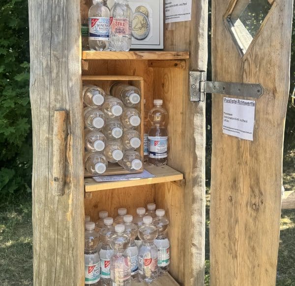 Holzschrank im Freien mit mehreren Flaschen Mineralwasser, Preisliste und Geldeinwurfbox für Selbstbedienung.