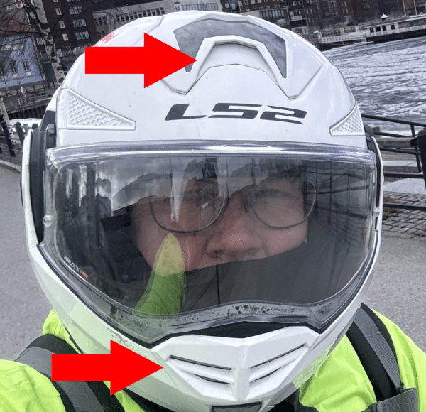 Person mit Brille trägt weißen Motorradhelm mit zwei roten Pfeilen, die auf Lüftungsschlitze zeigen. Hintergrund: Stadtgebäude am Wasser.