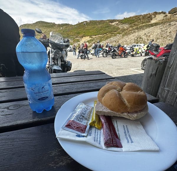 Ein Teller mit Brötchen und Saucenpäckchen, eine Wasserflasche auf Holztisch, im Hintergrund Motorräder und Menschen vor einer hügeligen Landschaft unter blauem Himmel.