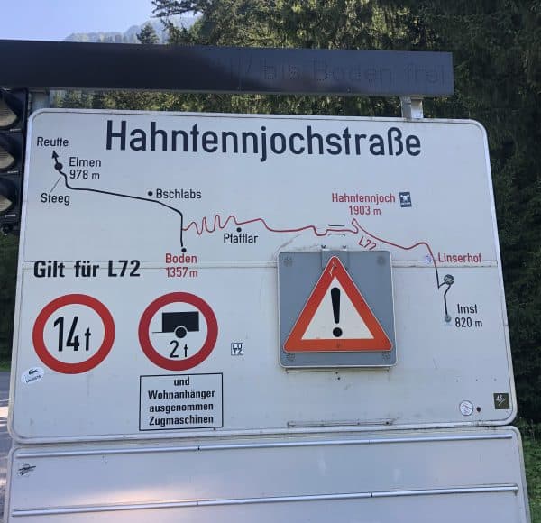 Straßenschild mit Karte der Hahntennjochstraße, Höhenangaben, Verkehrsbegrenzungen und Warnhinweisen, umgeben von Wald und Bergen im Hintergrund.