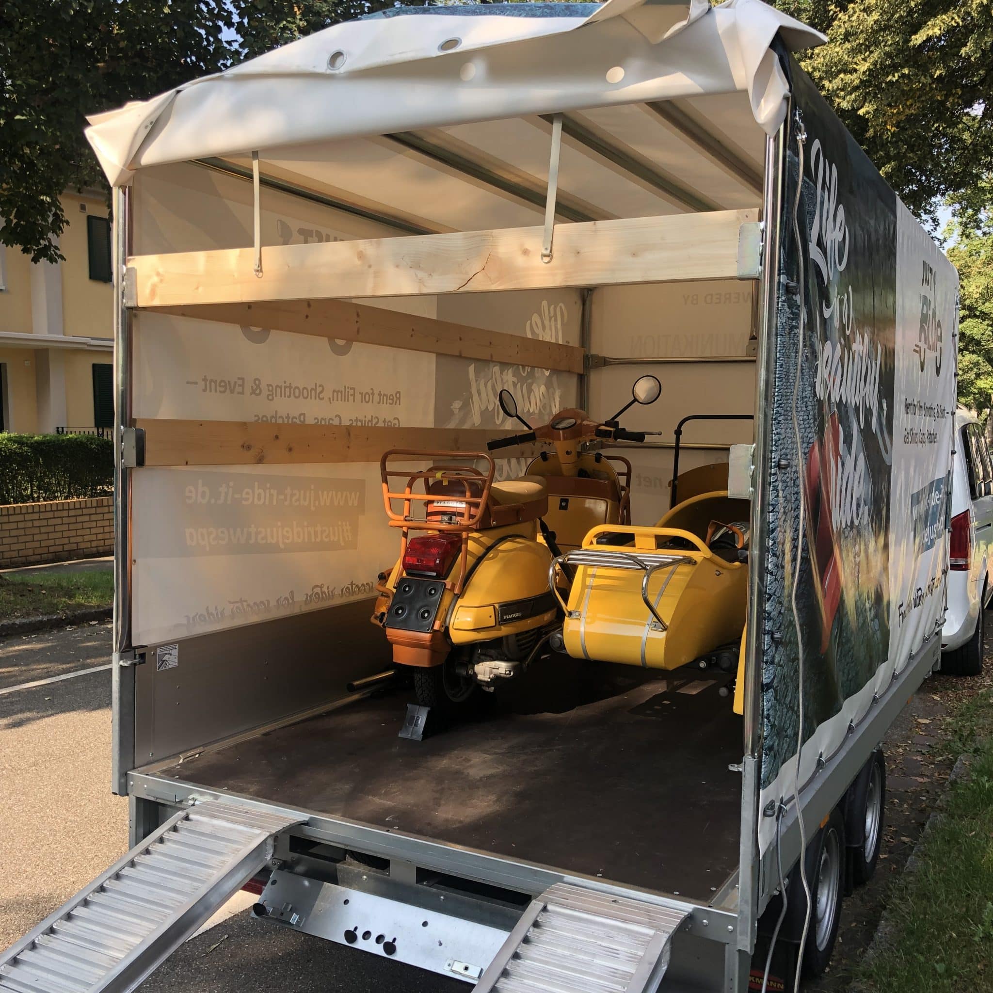 Zwei gelbe Motorroller mit Beiwagen stehen in einem offenen Transportanhänger auf einer Straße.