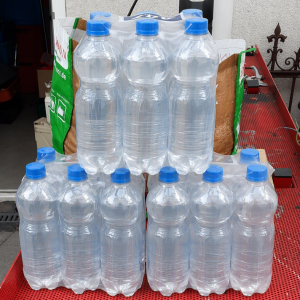 Mehrere Sixpacks mit Wasserflaschen aus Plastik, jeweils mit blauem Deckel, gestapelt auf einem roten Metalltisch im Freien.