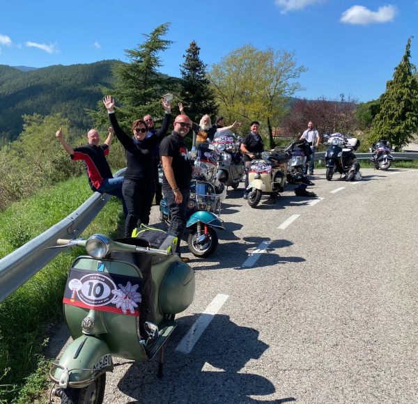 Eine Gruppe von Menschen mit Motorrollern posiert und winkt auf einer kurvigen Straße in einer grünen, hügeligen Landschaft bei sonnigem Wetter.