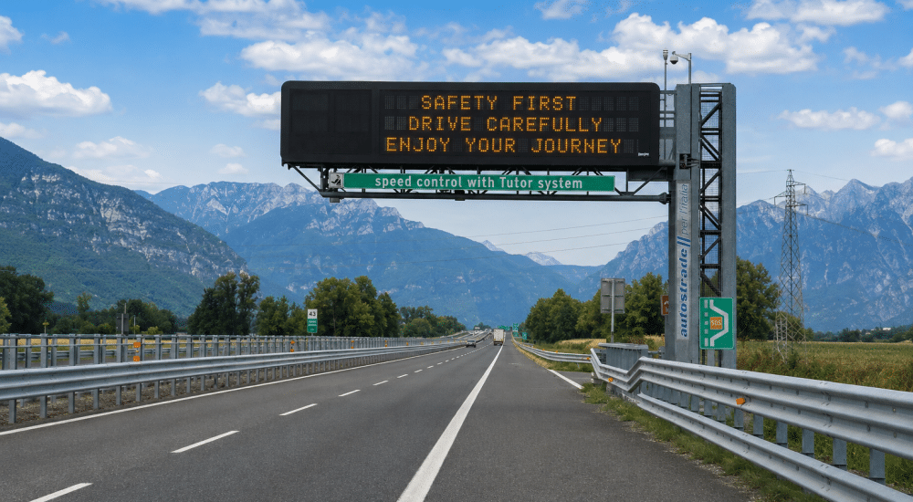 Autobahn mit elektronischem Verkehrsschild, das zur Vorsicht mahnt, vor Bergen und blauem Himmel, mit Leitplanken und SOS-Notrufstation am Straßenrand. © JustKommunikation GmbH | JustRide it