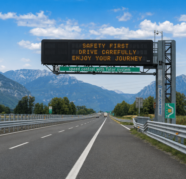 Autobahn mit elektronischem Verkehrsschild, das zur Vorsicht mahnt, vor Bergen und blauem Himmel, mit Leitplanken und SOS-Notrufstation am Straßenrand. © JustKommunikation GmbH | JustRide it