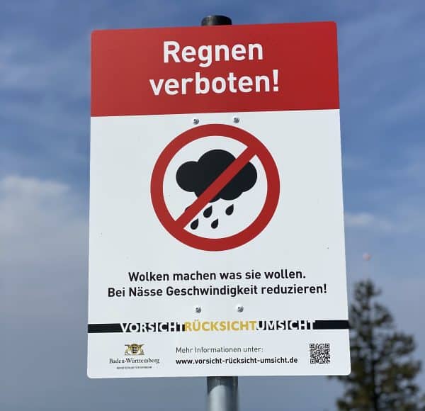 Verkehrsschild mit Aufschrift „Regnen verboten!“ und Symbol einer durchgestrichenen Regenwolke, darunter Hinweise zur Geschwindigkeit bei Nässe.